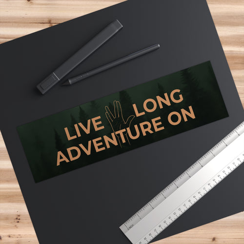 Black bumper sticker with 'Live Long Adventure On' text on a wooden surface with pens and a ruler.