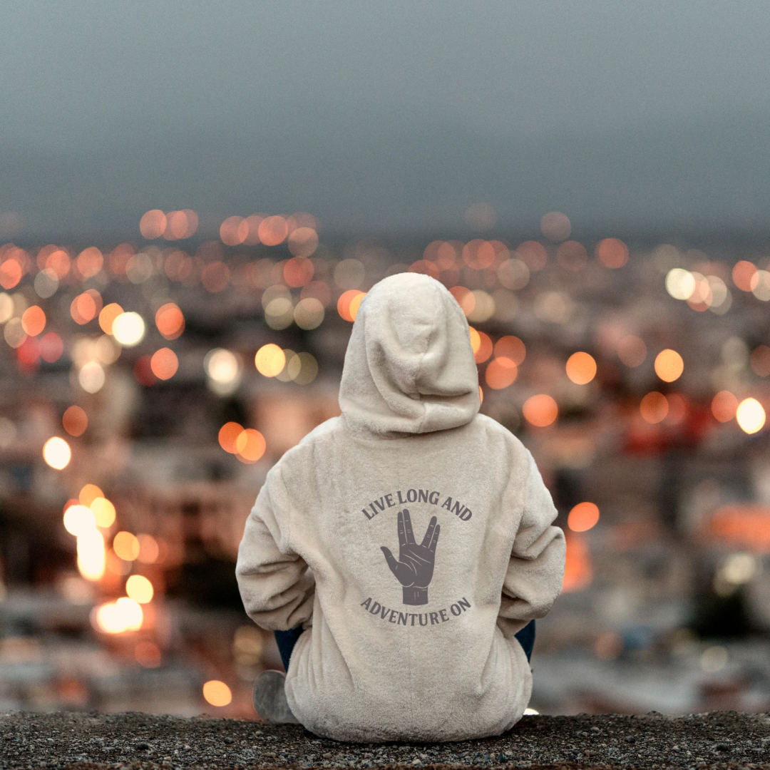 Person wearing a hoodie with text and a hand symbol, sitting on a rocky surface with a cityscape in the background.