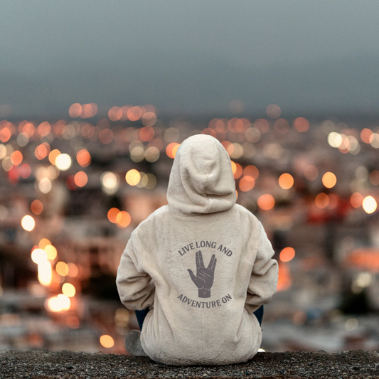 Person wearing a hoodie with text and a hand symbol, sitting on a rocky surface with a cityscape in the background.